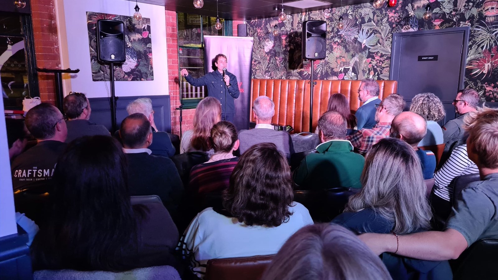 A comedian on stage with a microphone, performing to a packed room at a comedy night.